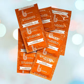 An assortment of bCondoms Studded Condoms, made from natural rubber latex and subjected to triple quality testing, is presented in orange packaging. The label "Studded" stands out alongside the message "Support Black Businesses," set against a white backdrop with a gentle bokeh effect.